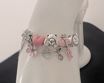 Op de afbeelding: Een zilveren armband met roze en zilveren bedels, waaronder een hartbedel met het woord "Moeder" erop, een lieveheersbeestje bedel, een teddybeer bedel, een bloemenbedel en een roze lintbedel. De armband heeft ook een bedel met het woord "Mama" erop.