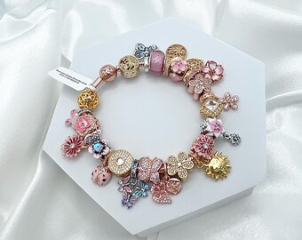 May include: A rose gold charm bracelet with various decorative charms. Charms include flowers, butterflies, a ladybug, and a sun. The bracelet is displayed on a white hexagonal stand against a white satin background.