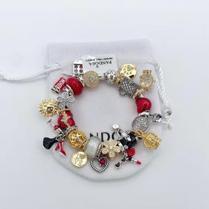 May include: A charm bracelet with various decorative charms, including red beads, gold crowns, and a lipstick charm. The bracelet is on a white drawstring bag with the word "Pandora" printed on it.