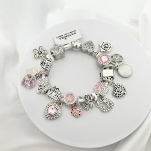 May include: A silver charm bracelet with various charms, including a flower, star, heart, and paw print. Some charms have pink accents and clear crystals. The bracelet is on a white plate.