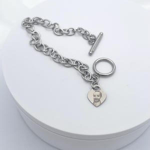 May include: A silver chain bracelet with a heart-shaped charm featuring a photo of a child. The bracelet has a toggle clasp.