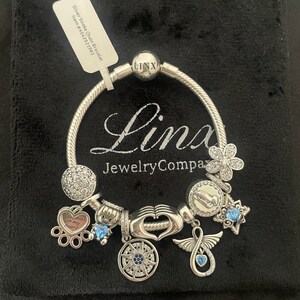 May include: Silver snake chain bracelet with charms, including a heart with the words "Paw prints on my heart", a hand with a heart, a compass, a star, a flower, a praying hands charm, and an angel wing charm. The bracelet is on a black velvet background.