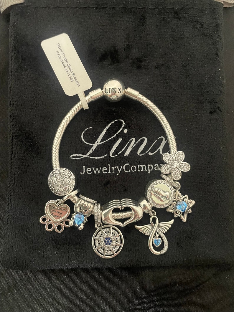 May include: Silver snake chain bracelet with charms, including a heart with the words "Paw prints on my heart", a hand with a heart, a compass, a star, a flower, a praying hands charm, and an angel wing charm. The bracelet is on a black velvet background.