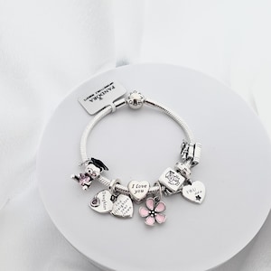 May include: A silver charm bracelet with multiple charms. Charms include a graduation bear, hearts with text, a pink flower, and a graduation cap. The bracelet is on a white surface.