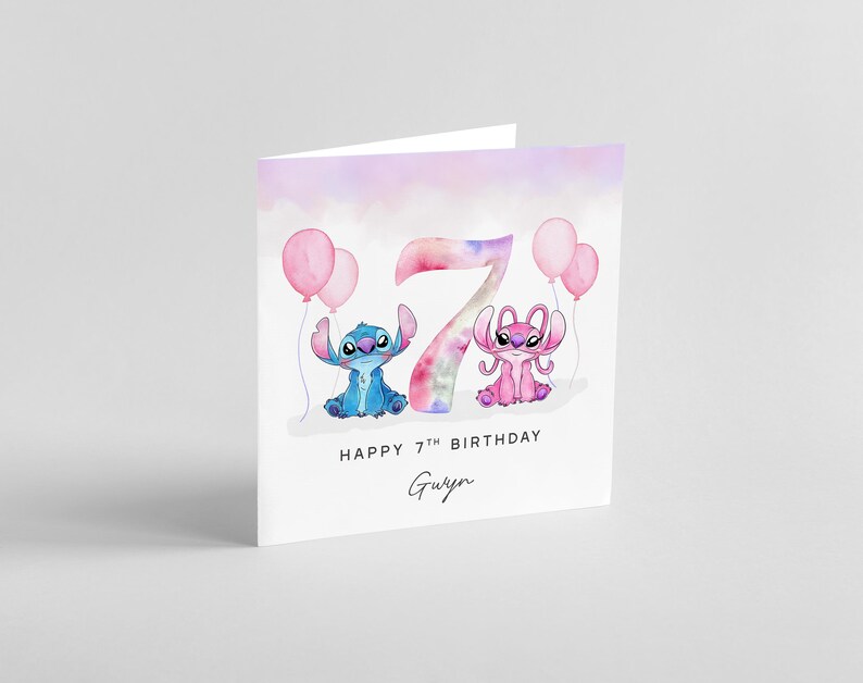 Peut inclure: Carte de v&oelig;ux blanche pli&eacute;e avec un motif aquarelle. La carte pr&eacute;sente un personnage de dessin anim&eacute; bleu et rose, des ballons roses et le chiffre 7. Le texte indique "HAPPY 7TH BIRTHDAY" et un nom en &eacute;criture manuscrite.