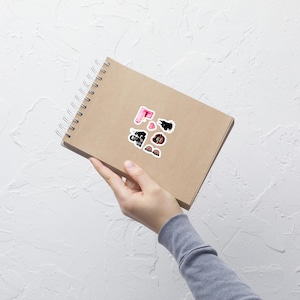 May include: A spiral-bound notebook with several stickers on the cover. The stickers include a pink lipstick, a black and white cow, a heart, and a pair of sunglasses. The notebook is held by a person.
