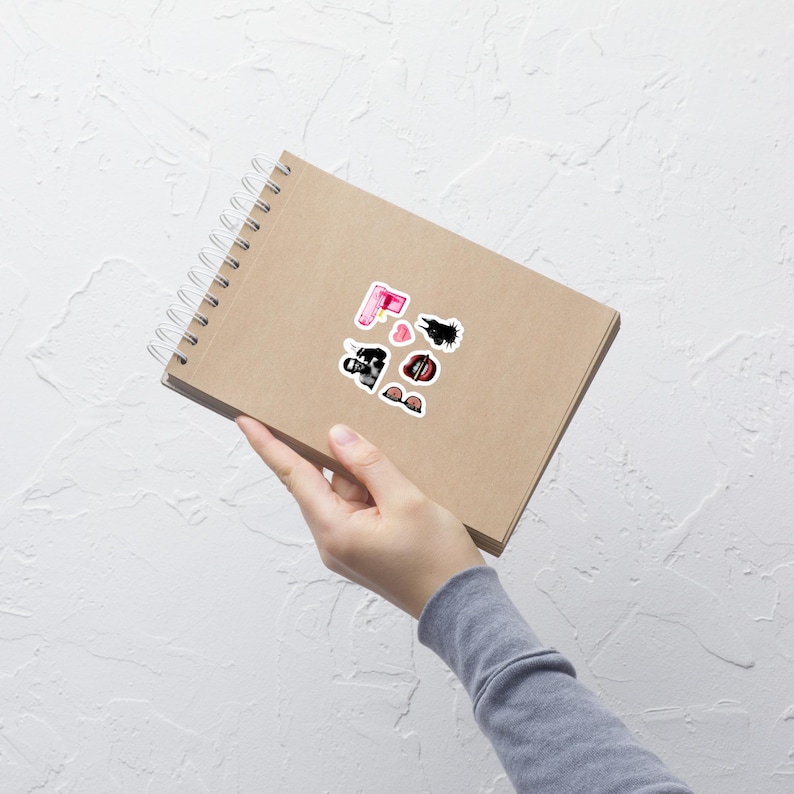 May include: A spiral-bound notebook with several stickers on the cover. The stickers include a pink lipstick, a black and white cow, a heart, and a pair of sunglasses. The notebook is held by a person.