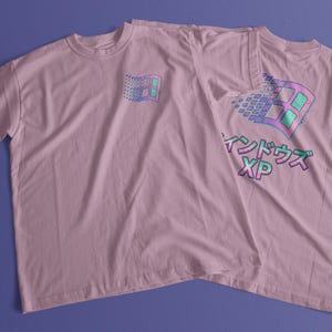 Op de afbeelding: Een lichtroze T-shirt met korte mouwen voorzien van een kleine afbeelding van een gestileerd computervenster op de linkerborst en een grotere versie van hetzelfde ontwerp op de achterkant met de Japanse tekst "ウィンドウズ" en de letters "XP" in verloop.