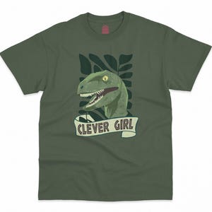 Può includere: Maglietta verde con una grafica di una testa di dinosauro e il testo "Clever Girl".