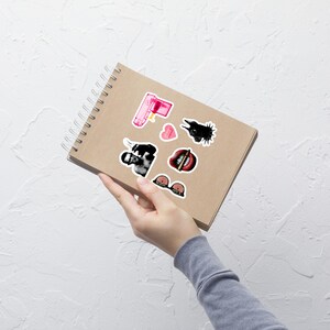 May include: A spiral-bound notebook with several stickers. The stickers include a pink water gun, a black and white image of a person, a heart, a black bird, a mouth with a bullet, and a bra.