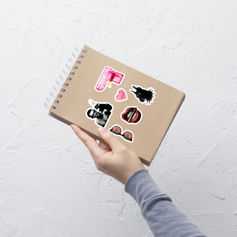 May include: A spiral-bound notebook with several stickers. The stickers include a pink water gun, a black and white image of a person, a heart, a black bird, a mouth with a bullet, and a bra.