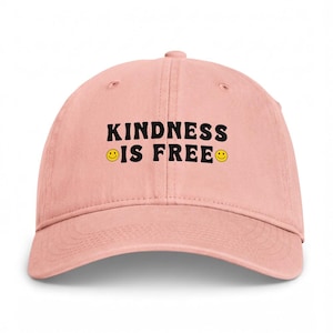 Può includere: Un cappellino da baseball rosa chiaro con la scritta nera "KINDNESS IS FREE" stampata sul davanti. Il design include due piccole faccine sorridenti gialle, una posizionata prima della parola "IS" e una dopo la parola "FREE" sul cappellino.
