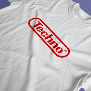 May include: A white short-sleeved t-shirt featuring a red graphic on the chest. The graphic consists of the word Techno written in a bold, sans-serif font, enclosed within a red oval outline that is tilted at an angle. A small registered trademark symbol is visible.