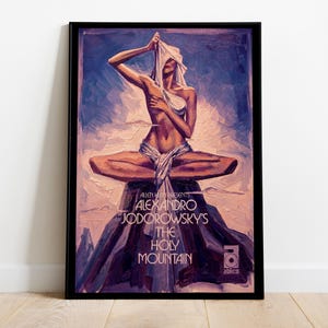 May include: A framed art print featuring a figure in a meditative pose atop a mountain. The artwork, in shades of blue, purple, and brown, includes the text "ALEXANDRO JODOROWSKY'S THE HOLY MOUNTAIN" and the Allen Klein Presents logo.