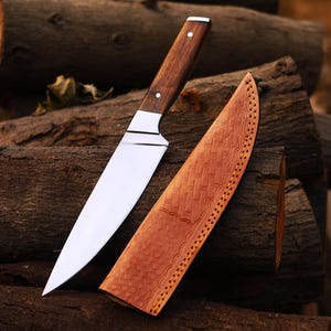May include: A stainless steel chef's knife with a dark wood handle and a leather sheath. The knife blade is long and sharp, and the sheath is a light brown color with decorative stitching. The knife and sheath are resting on a pile of wood logs.