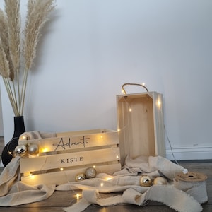 May include: Two wooden crates, one with the word "Advents Kiste" and the other empty, are illuminated by string lights. Gold ornaments and a beige fabric add to the festive display. A black vase with tall, dried grass is in the background.