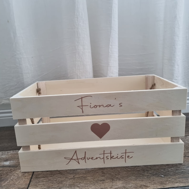 May include: A light-colored wooden crate with the text "Fiona's Adventskiste" in a script font. A brown heart is painted on the crate. The crate is made of wooden slats and has rope handles.
