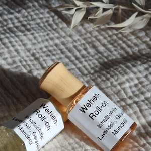 Könnte beinhalten: Zwei Roll-on-Flaschen mit Holzverschlüssen und Etiketten. Die Etiketten tragen die Aufschrift "Wehen-Roll-on" und listen Inhaltsstoffe wie Lavendel, Geranie und Mandelöl auf. Die Flaschen stehen auf einer strukturierten, cremefarbenen Oberfläche.