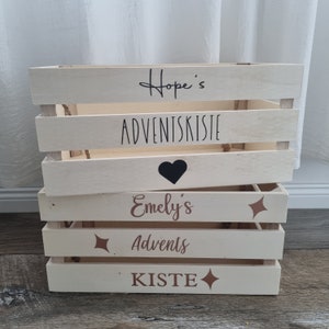 May include: A stack of four light-colored wooden crates. The top crate has the text "Hope's ADVENTSKISTE" in black script. The second crate has a black heart. The third crate has the text "Emely's Advents KISTE" in brown script with starburst accents.