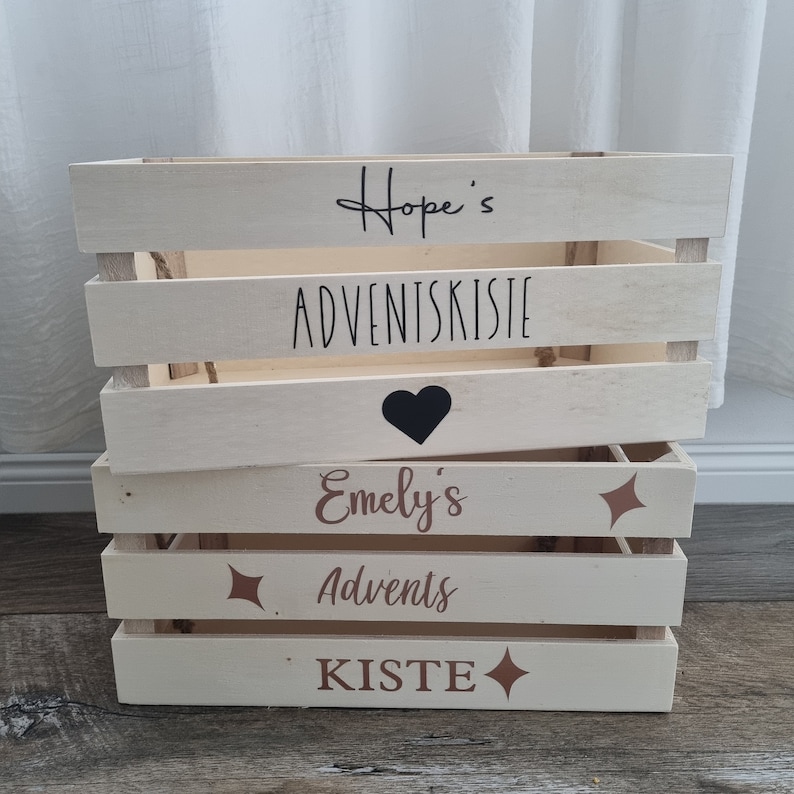 May include: A stack of four light-colored wooden crates. The top crate has the text "Hope's ADVENTSKISTE" in black script. The second crate has a black heart. The third crate has the text "Emely's Advents KISTE" in brown script with starburst accents.
