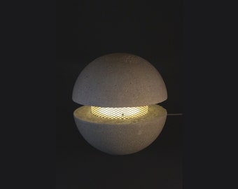 May include: A gray concrete table lamp with a wire mesh shade. The lamp is turned on and the light shines through the mesh.