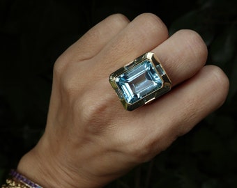 May include: A gold ring with a large, rectangular, light blue gemstone. The ring is on a person's finger.