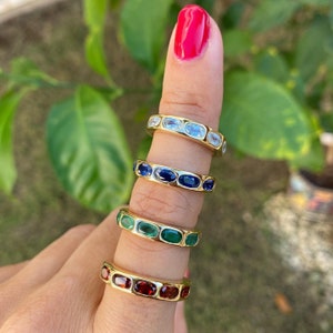 May include: Four gold rings with different coloured gemstones. The rings are stacked on a finger, with the top ring featuring clear gemstones, followed by blue, green, and red gemstones.