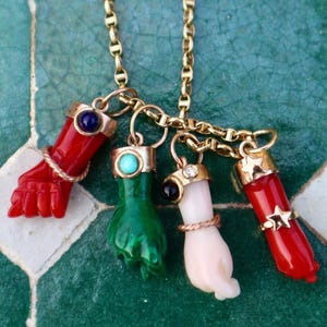 Puede incluir: Un collar dorado con cuatro colgantes en forma de mano en rojo, verde, blanco y rojo. Cada colgante tiene un anillo y está adornado con piedras preciosas. El collar se exhibe sobre una superficie de baldosas verdes y blancas.