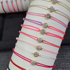 May include: A display of colourful string bracelets, each with a single, sparkling, round diamond set in a gold bezel. The bracelets are presented on a white cylindrical form, showcasing a variety of colours.