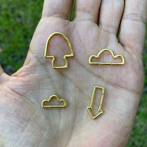 Può includere: Quattro portachiavi in metallo dorato di diverse forme: un fungo, due nuvole e una freccia. I portachiavi sono mostrati su una mano.