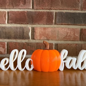 May include: A white 3D printed sign with the text hello fall in a cursive script font, featuring a bright orange pumpkin with a brown stem positioned in the center of the word hello and the word fall, creating a single decorative display.