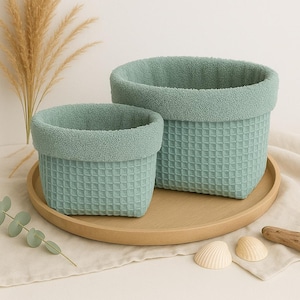 Könnte beinhalten: Zwei passende aqua-farbene Aufbewahrungskörbe mit Waffelmuster. Die Körbe haben einen weichen, flauschigen Besatz am oberen Rand. Sie stehen auf einem runden, hellbraunen Holztablett, mit dekorativen Muscheln und einem Holzstück.