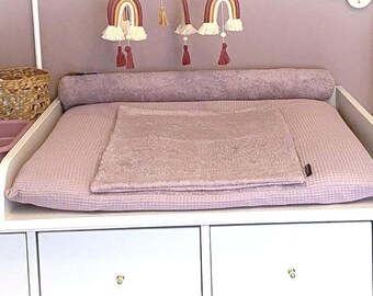 May include: A mauve waffle-textured changing pad with a matching bolster pillow and a rectangular coverlet. The set is placed on a white dresser alongside a mauve plastic basin, a woven basket, and a cylindrical lamp with a rattan shade.