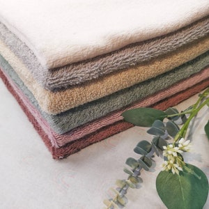 Könnte beinhalten: Ein Stapel weicher, flauschiger Handtücher in verschiedenen gedämpften Farben, darunter Weiß, Grau, Beige und Altrosa. Die Handtücher sind ordentlich gefaltet, mit einem Zweig Eukalyptus und weißen Blüten in der Ecke.