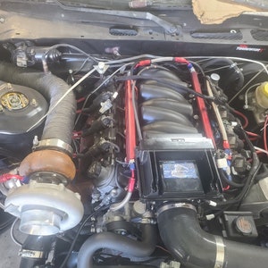 Puede incluir: Vista detallada de un motor de coche, que muestra un turbocompresor, rieles de combustible rojos y varias mangueras y cables. El motor está parcialmente ensamblado, con un enfoque en las modificaciones de rendimiento y los componentes personalizados.