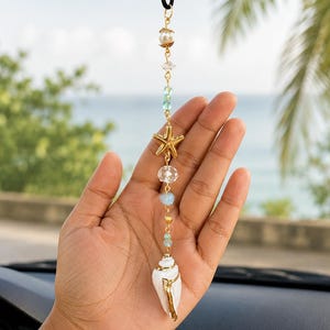 May include: A hanging car ornament featuring a white seashell with gold-tone trim, a gold-tone starfish charm, and a vertical arrangement of clear, light blue, and pearl-like beads on a gold-tone chain suspended from a black cord.