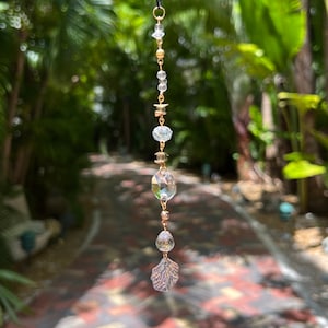 May include: A hanging suncatcher with a black cord and gold-colored metal accents. The suncatcher features clear, faceted crystal beads and a decorative, iridescent seashell charm. The background is a blurred view of lush green foliage and a brick pathway.