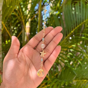 May include: A gold-colored pendant necklace featuring a clear, faceted crystal at the bottom, with a starfish charm and various beads in shades of blue, clear, and gold. The necklace is suspended from a black cord, against a backdrop of green palm leaves.