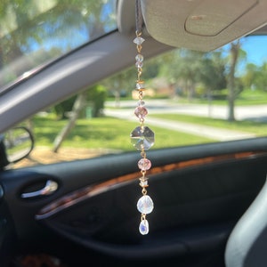 May include: A car charm featuring a string of clear and pink crystals, with a large faceted crystal at the center. The charm is suspended from a black string and hangs in front of a car's interior.