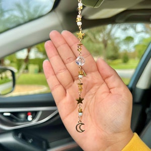 May include: A gold car charm with a crescent moon and stars hanging from a rearview mirror. The charm is made of beads and metal.