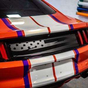May include: The back of a red car with white and purple racing stripes. A black and white American flag decal is on the rear window.