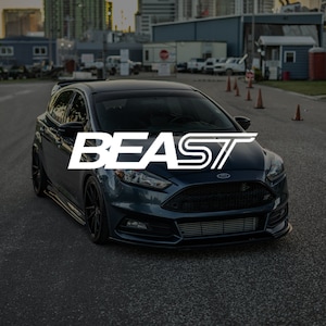 May include: A dark blue Ford Focus ST with the word "BEAST" in white letters across the front of the car. The car is parked on a black tarmac surface.