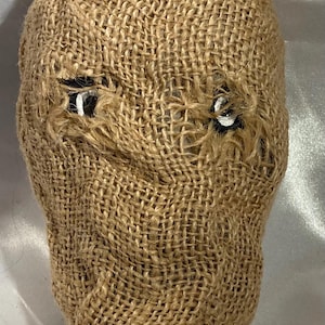 Puede incluir: Una máscara de saco de arpillera marrón con dos ojos blancos pintados. La máscara tiene una textura rugosa y está atada en la parte superior.