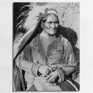 May include: A black and white portrait of a Native American man wearing a feathered headdress and a fringed jacket. He is holding a revolver in his lap.