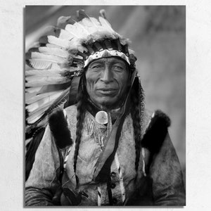 May include: A black and white portrait of a Native American man wearing a traditional headdress with feathers. He is looking directly at the camera with a serious expression.