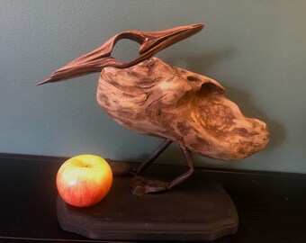 Peut inclure: Une sculpture d'oiseau en bois avec un long bec incurvé et une surface rugueuse et texturée. L'oiseau est perché sur un support en métal et a une petite pomme rouge devant lui.