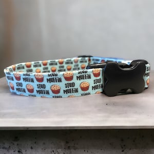 May include: A light blue dog collar with a black plastic buckle. The collar has a repeating pattern of muffins with the text "Stud Muffin" on each muffin.