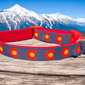 Könnte beinhalten: Ein rotes und graues Hundehalsband mit dem Design der Colorado-Staatsflagge. Das Halsband hat eine rote Basis mit einem grauen Band, das das Colorado-Flaggensymbol in Rot und Gelb zeigt. Ein goldfarbener Metallring und ein roter Kunststoffverschluss sind sichtbar.