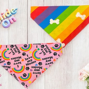 May include: Two dog bandanas. One is rainbow-colored with white bone shapes. The other is pink with rainbows, paw prints, and the text "I love my Moms". The bandanas are on a light wood-grain surface.
