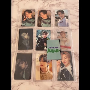 May include: A collection of photo cards featuring different members of a K-pop group. The cards are arranged in a clear plastic binder with a green and purple sticker that says "KPOPPIN' MANIAC".